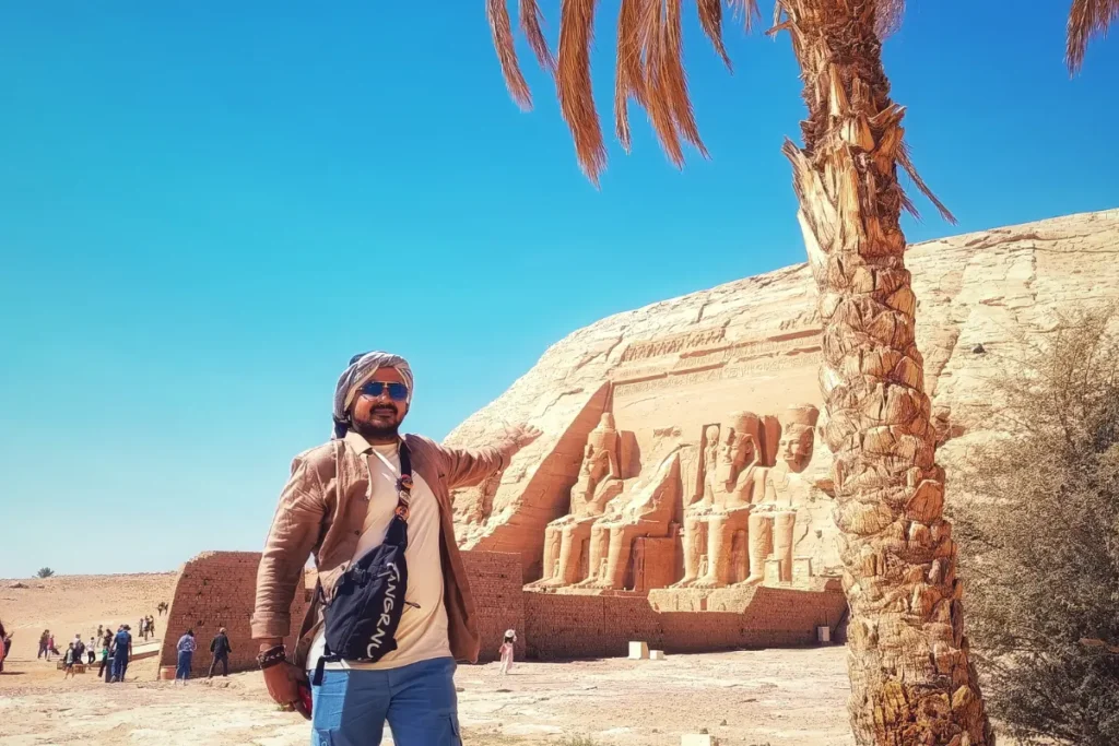 Abu Simbel Akash wearing a white shirt, sunglasses, and a headscarf poses proudly on a sunny day in front of the massive stone statues at the Abu Simbel temple.