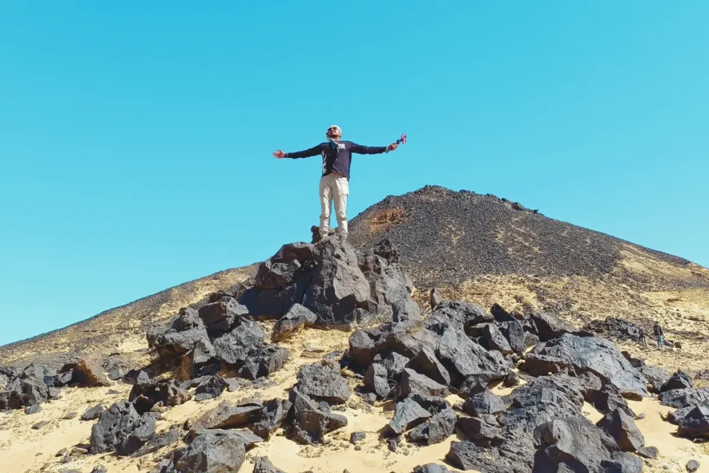 Akash standing on top of a dark, volcanic hill at the Black Desert in Egypt, overlooking a vast desert landscape dotted with black-topped volcanic mounds.