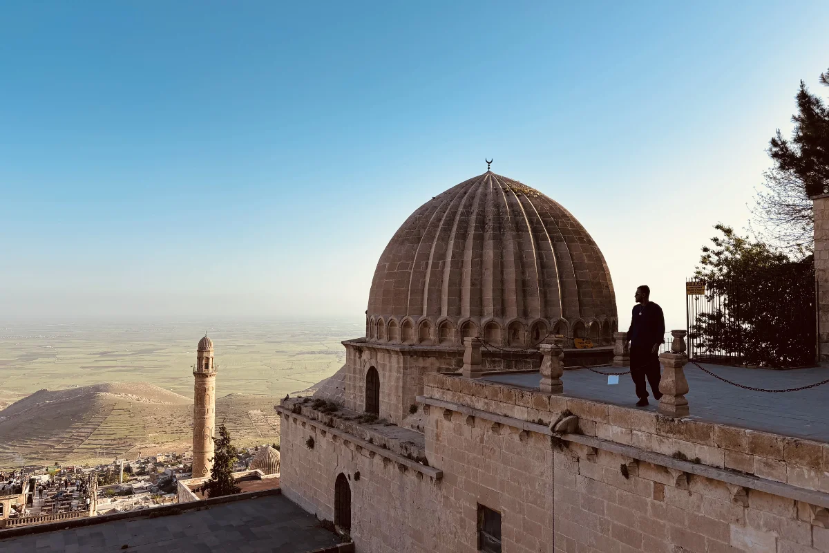 Silhouette of Akash standing on a rooftop balcony of Zincriye Madrasa next to a large dome structure, overlooking a vast, misty landscape during twilight.