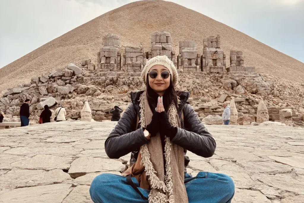 Sumana sitting cross-legged on the West Terrace of Mount Nemrut with giant stone heads scattered around her against a cloudy sky.