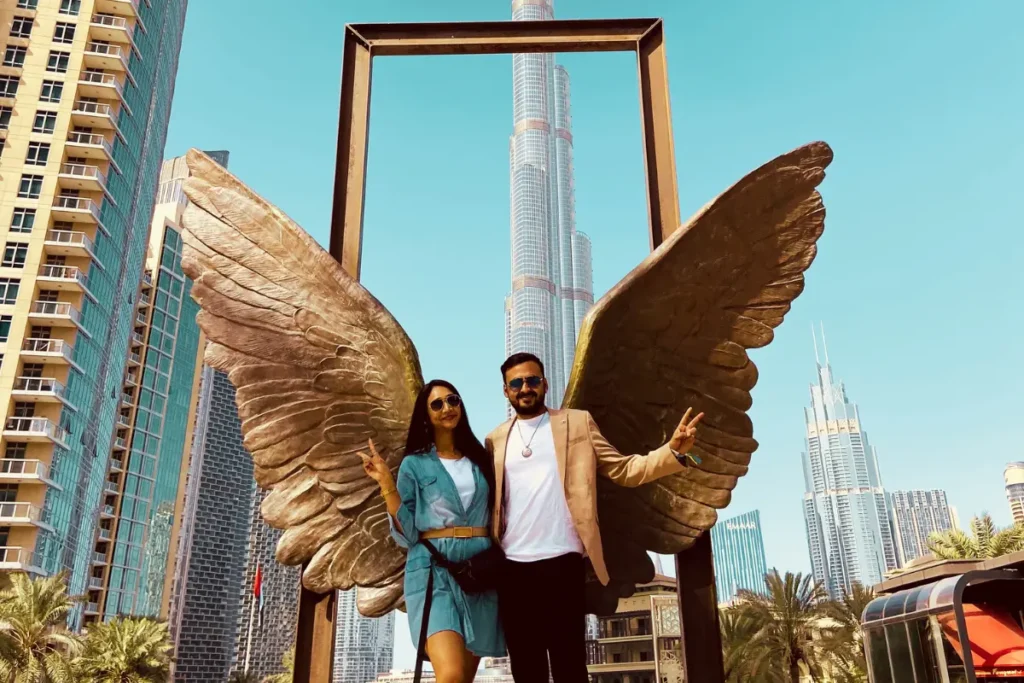 Akash & Sumana posing on a high-altitude glass platform with large, artistic golden wings spread open behind them. Backdrop of Wings of Mexico