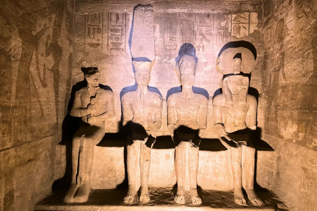 A dimly lit stone chamber inside the heart of an ancient Egyptian temple, showing four stone statues seated side-by-side against the back wall.