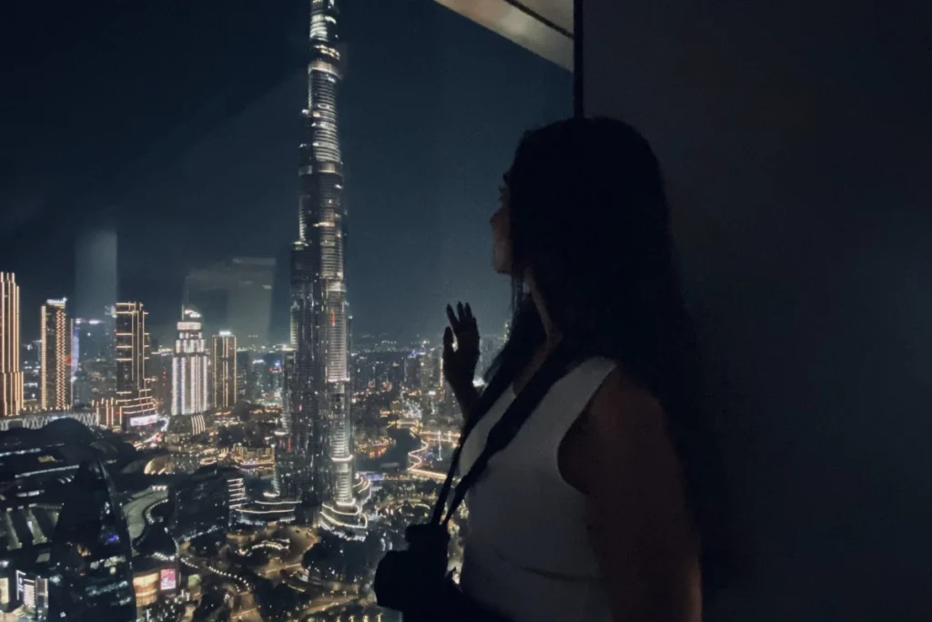Sumana standing against a glass window, looking out over the glowing night skyline of Dubai and the towering Burj Khalifa.