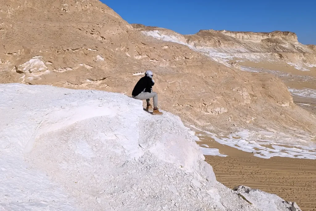 Akash wearing a black tshirt sitting on the edge of a high, bright white limestone cliff overlooking a desert valley.