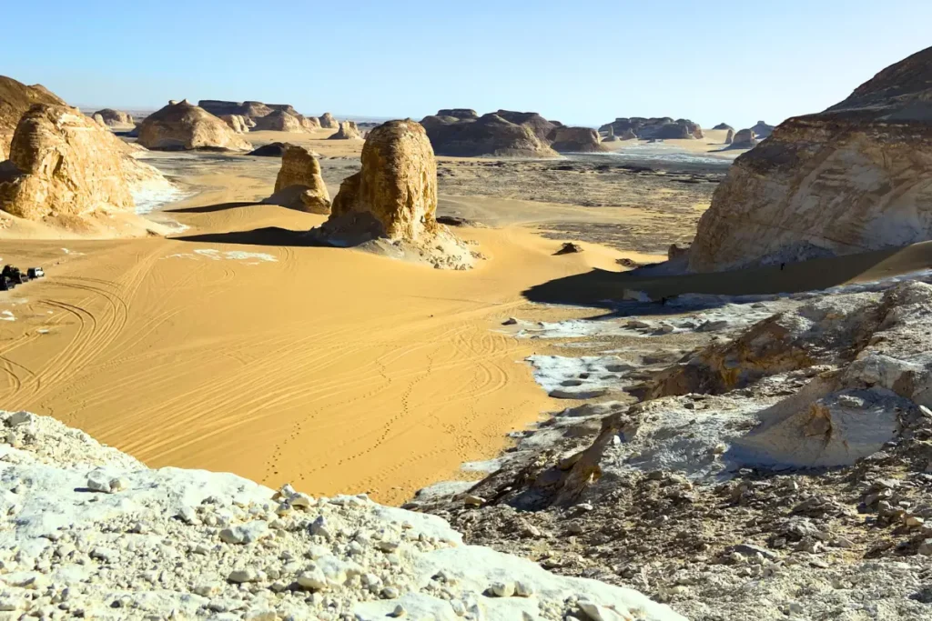 A scenic view of the Agabat Valley showing golden sand dunes interspersed with white chalk limestone formations under a clear blue sky.