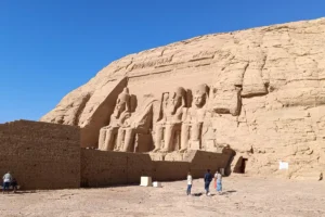 A wide landscape photograph of the massive rock-cut sandstone facade of the Great Temple of Abu Simbel under a clear blue sky, featuring four immense seated statues of Pharaoh Ramesses II.