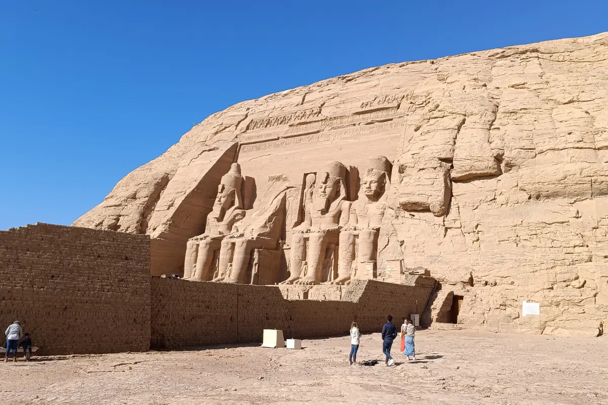 A wide landscape photograph of the massive rock-cut sandstone facade of the Great Temple of Abu Simbel under a clear blue sky, featuring four immense seated statues of Pharaoh Ramesses II.