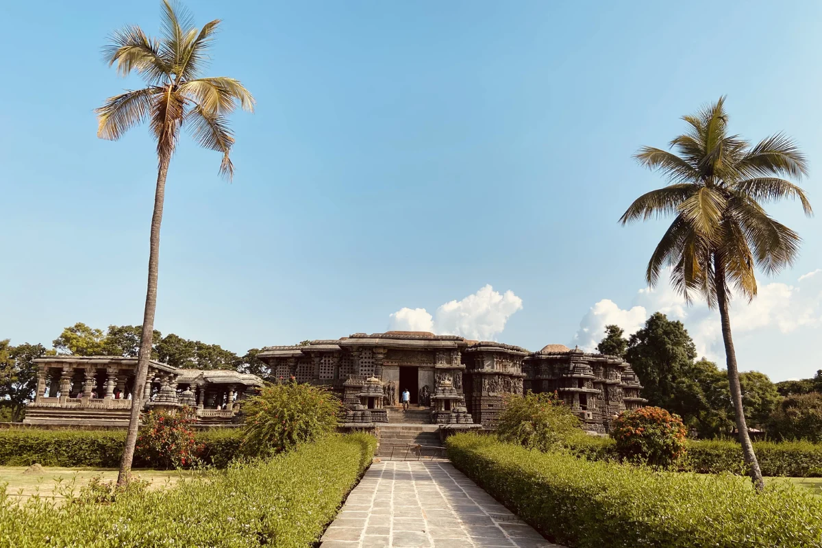 A wide exterior shot of the star-shaped Hoysaleshwara Temple featuring detailed stone carvings along its walls and pillars, flanked by tall palm trees under a bright blue sky.