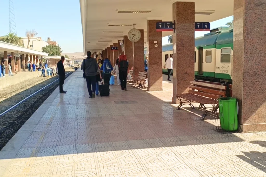 Travelers walking along an open-air platform under geometric metal canopies at the Aswan train station.