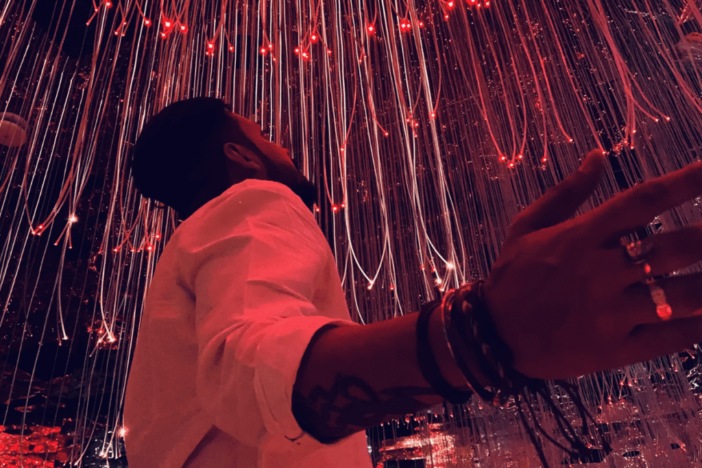Akash standing in an immersive digital art room with vertical strings of neon blue and yellow lights cascading from the ceiling.