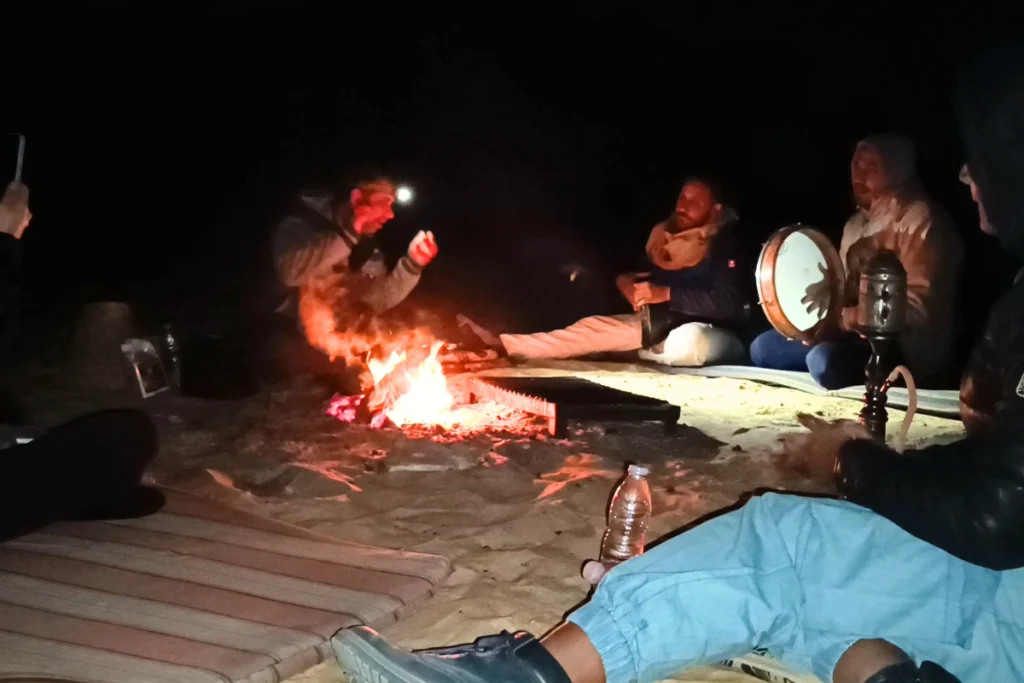 A group of people sitting in a circle on blankets around a glowing campfire in the pitch-black desert night.