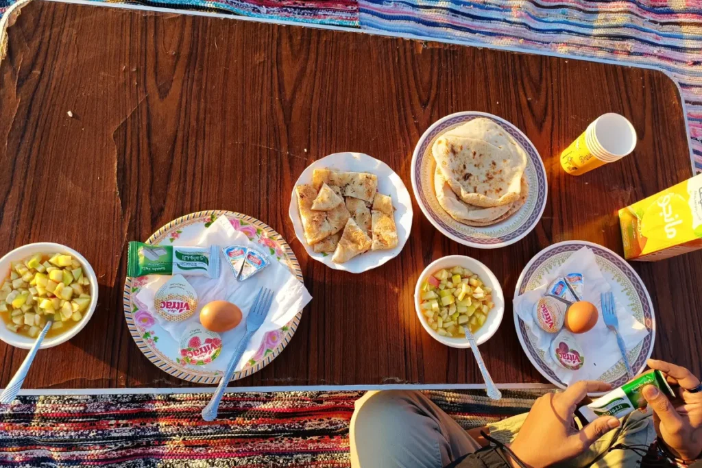A colorful breakfast spread featuring eggs, fresh fruits, vegetables, and bread served on a blanket over the white sand.