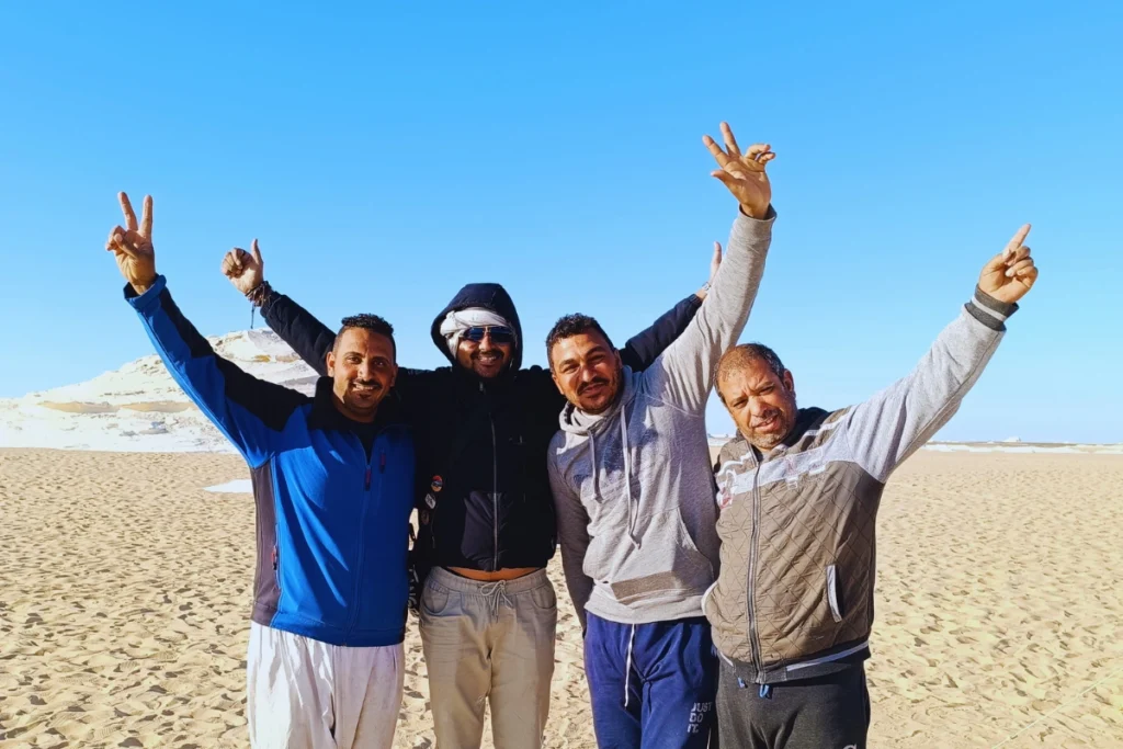 A group of four cheerful men posing shoulder-to-shoulder with peace signs against a clear blue desert sky.