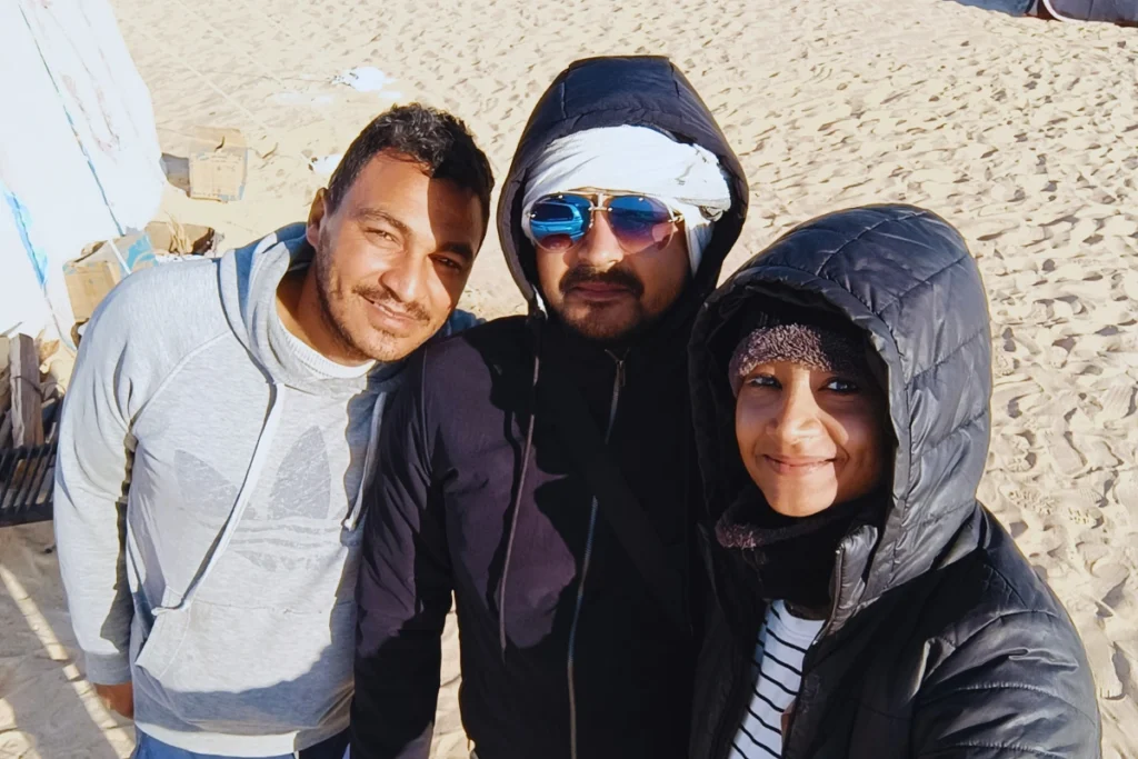 A group selfie of Akash & Sumana smiling together, bundled in winter gear against a bright, sunny backdrop.