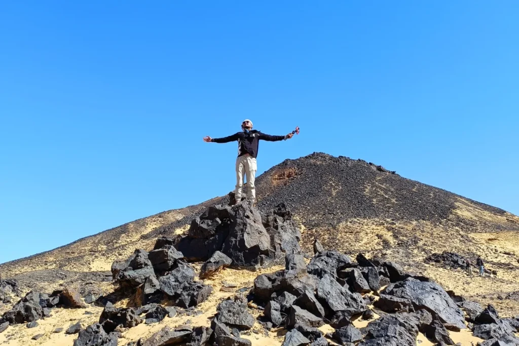 Akash with his arms outstretched stands on a dark, volcanic stone outcrop looking over black-topped hills in Egypt.