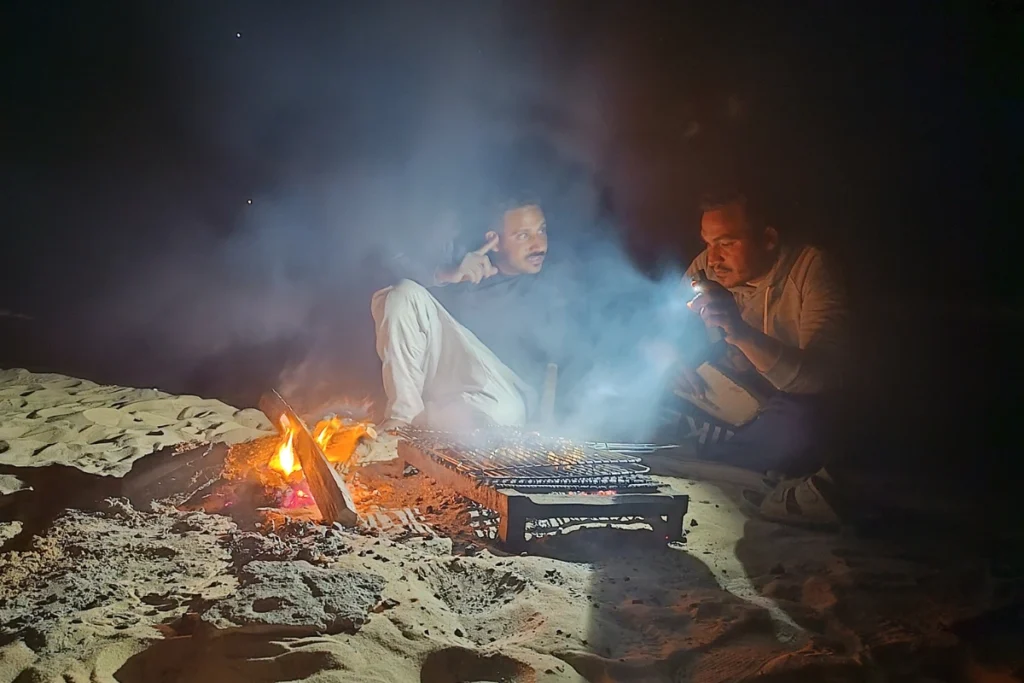 A close-up view of a guide kneeling by a campfire, grilling meat skewers under the dark night sky.