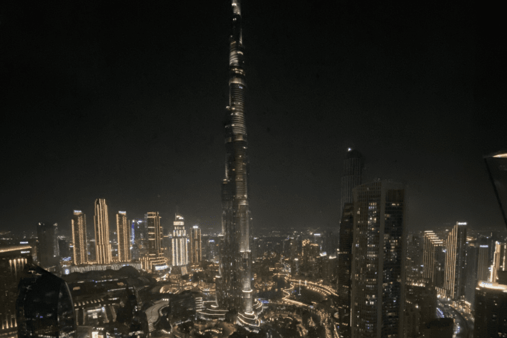 A nighttime view from an elevated viewpoint looking across the brightly lit cityscape toward the illuminated Burj Khalifa skyscraper.