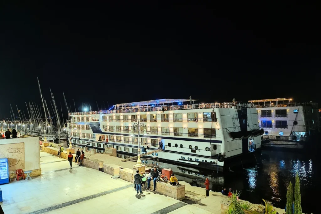 A large white cruise liner lit up at night, moored along a paved promenade where people are walking.