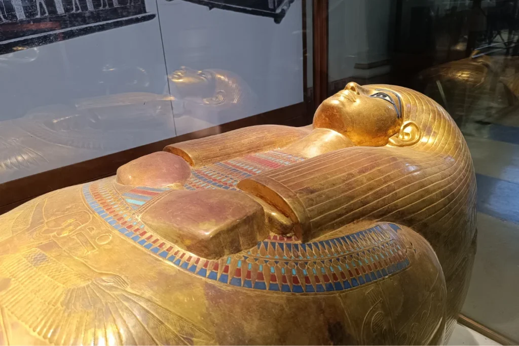 A side-angle close-up of a lavishly gilded ancient Egyptian anthropoid coffin covered in gleaming gold leaf inside a museum glass display case.