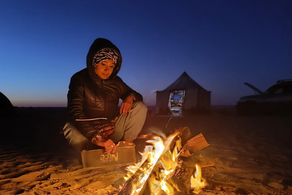 A young woman wearing a warm dark jacket and a maroon knit beanie sitting cross-legged on a patterned blanket on the sand, smiling next to a glowing wood campfire in the desert at night.