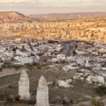 An aerial perspective of Göreme displaying white stone buildings, cave dwellings, and tall fairy chimney rock structures under warm sunlight.