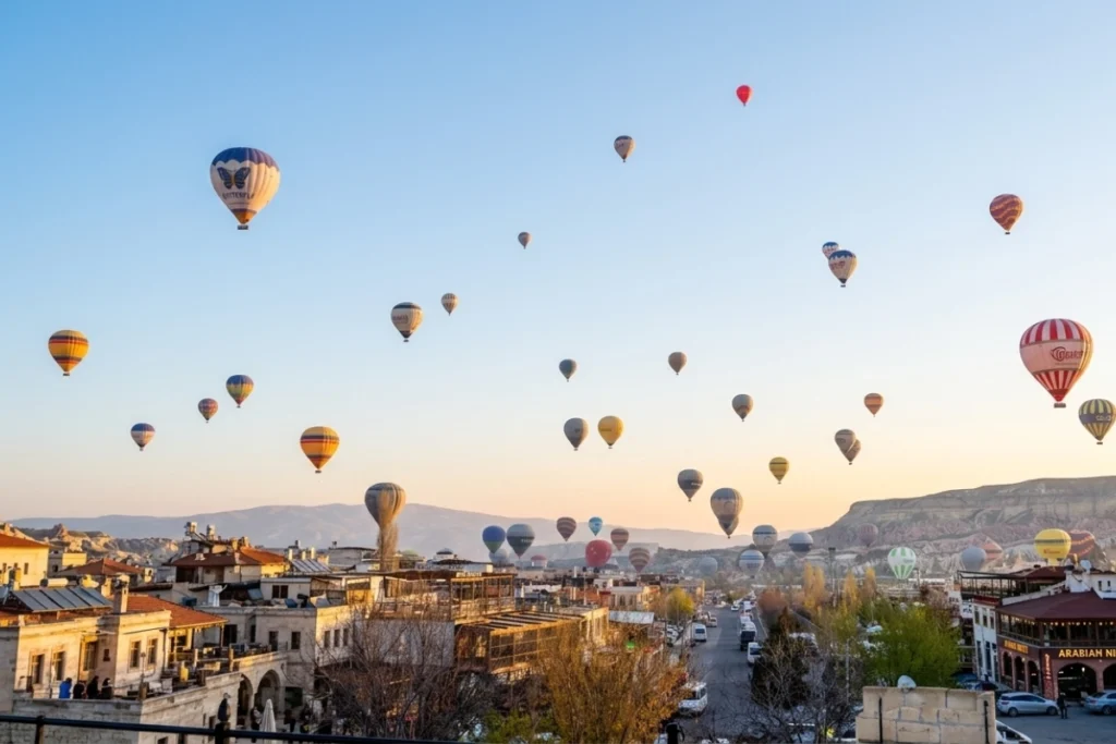 Cappadocia Guide banner (1) Dozens of colorful hot air balloons soaring over the ancient cave buildings and rock formations of Cappadocia at sunrise.