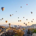 A wide panoramic landscape shot of Cappadocia at dawn, with numerous colorful hot air balloons dotting the soft sky above rock-cut houses and unique rock formations.