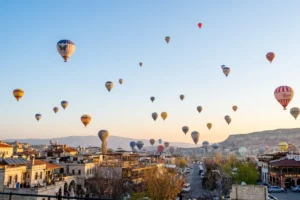 A wide panoramic landscape shot of Cappadocia at dawn, with numerous colorful hot air balloons dotting the soft sky above rock-cut houses and unique rock formations.