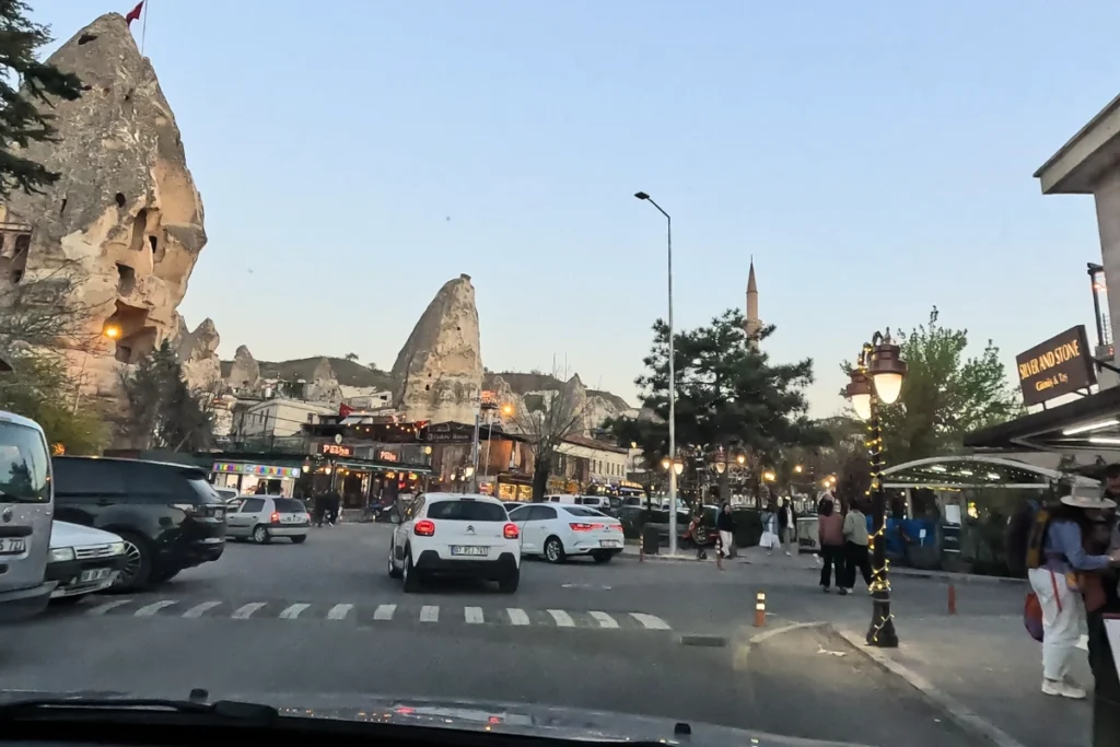 A bustling street scene in Göreme town center with shops, cars, and a massive natural rock formation towering over the buildings.