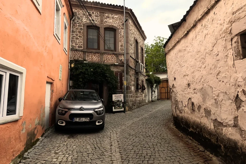 A narrow, historic cobblestone village street flanked by traditional colorful houses, with a car parked outside.