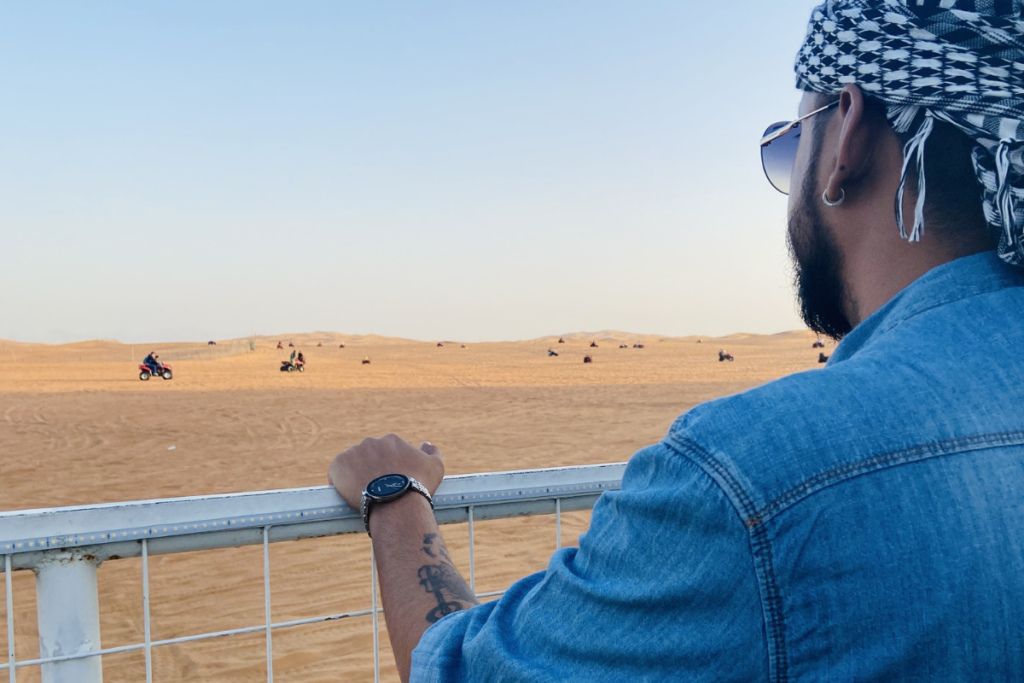Akash posing on a sandy desert dune while dressed in a traditional white Kandura and a checkered blue-and-white Ghutrah headdress.