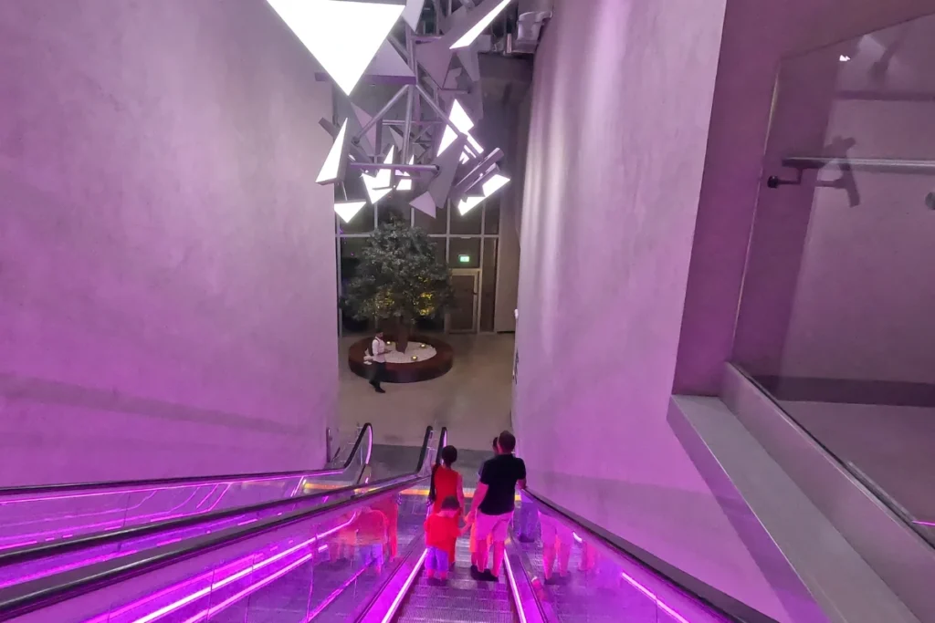 A wide view of a modern building interior featuring an ornate, illuminated circular elevator shafts structure surrounded by indoor landscaping.