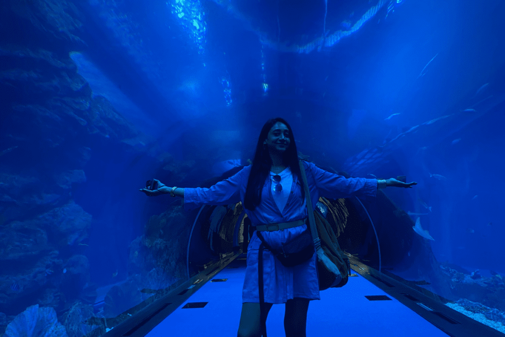 Sumana posing under a massive underwater glass tunnel inside the Dubai Mall Aquarium, with large fish swimming overhead in deep blue water.