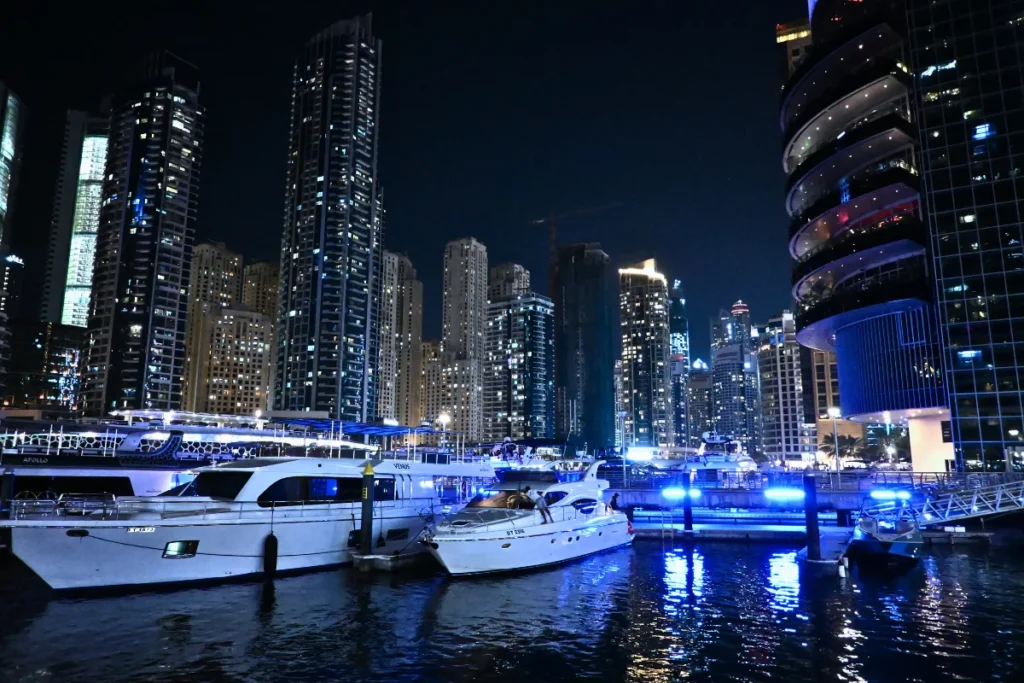 A panoramic view of the Dubai Marina waterway at night, filled with illuminated yachts and bordered by glowing modern high-rises.