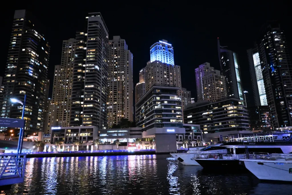 A vivid nighttime view across the water at Dubai Marina, reflecting the bright blue and purple neon lights of the surrounding luxury skyscrapers.