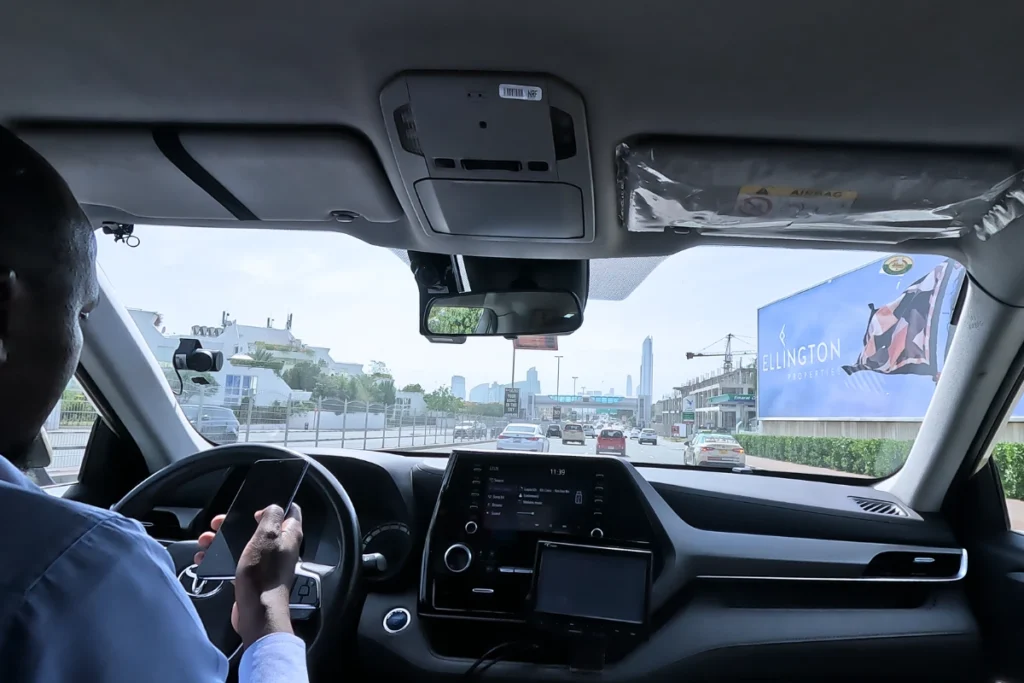 A point-of-view perspective looking past a driver's shoulder at a busy multilane highway stretching through urban architecture.