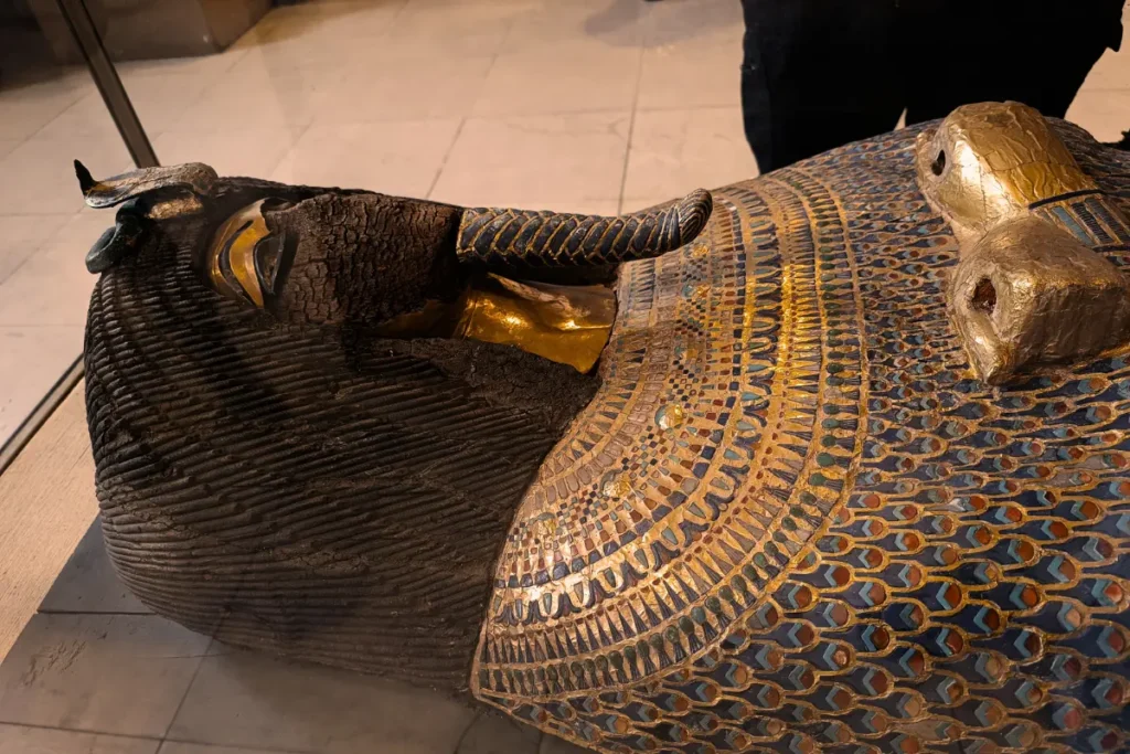 An ancient Egyptian anthropoid coffin with a dark black resin finish, adorned with crossing bands of bright gold leaf inscribed with hieroglyphs.