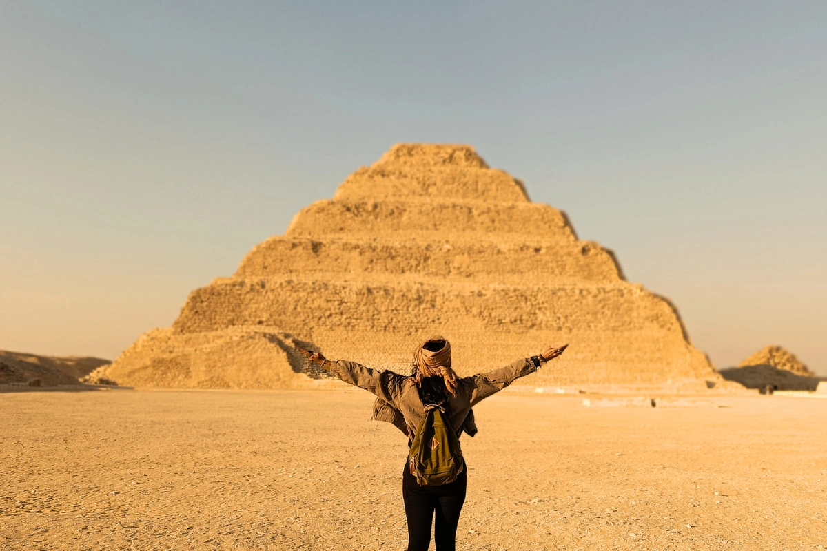Sumana with arms outstretched stands in the foreground, facing the historic stone tiers of the Step Pyramid in Saqqara under a clear blue sky.