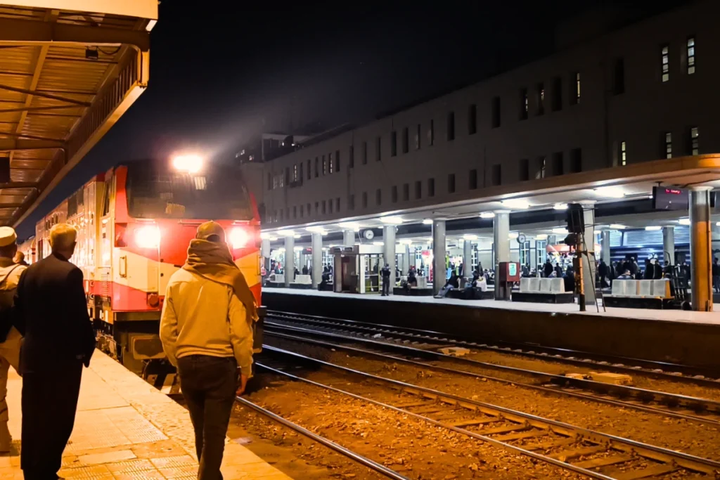 A side-angle shot of commuters standing on a train platform next to a stationary orange and white locomotive under station lights.