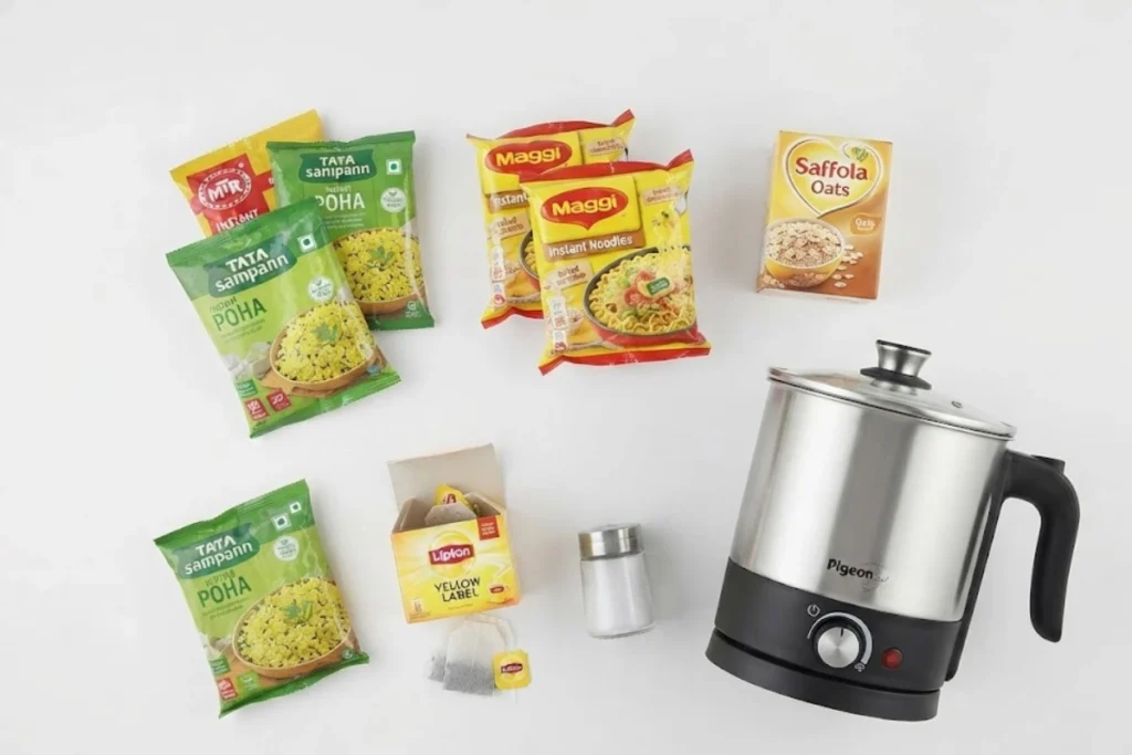 A collection of instant noodle packets, seasoning sachets, a small salt shaker, and a compact silver-and-black electric travel kettle set up on a surface.
