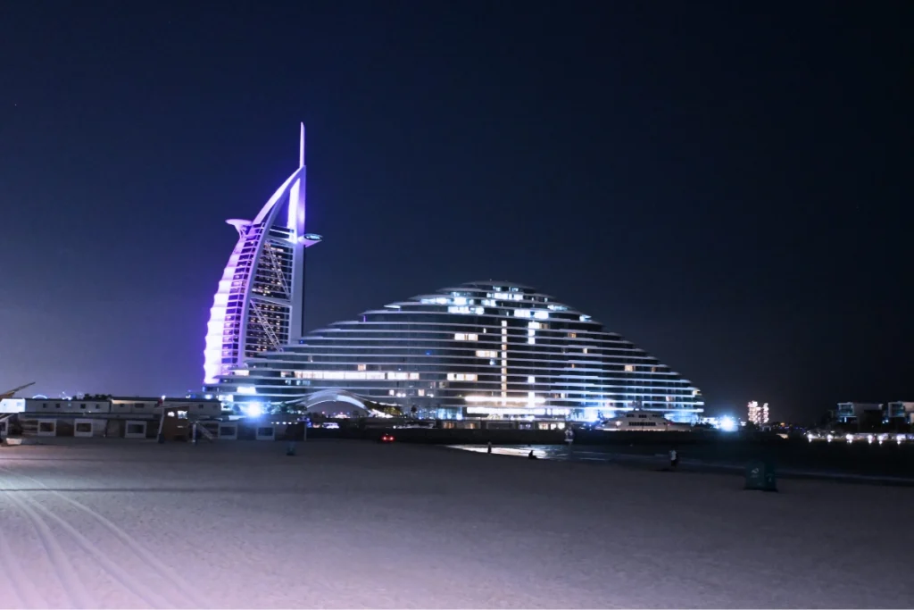 A wide view from a sandy beach looking across the water toward the sail-shaped architectural structure of the Burj Al Arab illuminated at dusk.