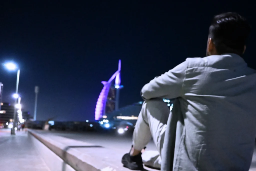 Akash sitting quietly on the sand at night, looking out across the calm dark water toward the brightly lit Burj Al Arab hotel.
