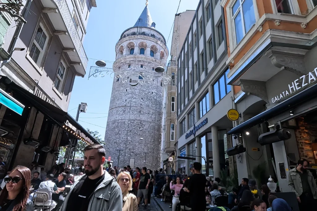 Looking up from a narrow city street at the towering stone structure of the historic Galata Tower reaching into the sky.