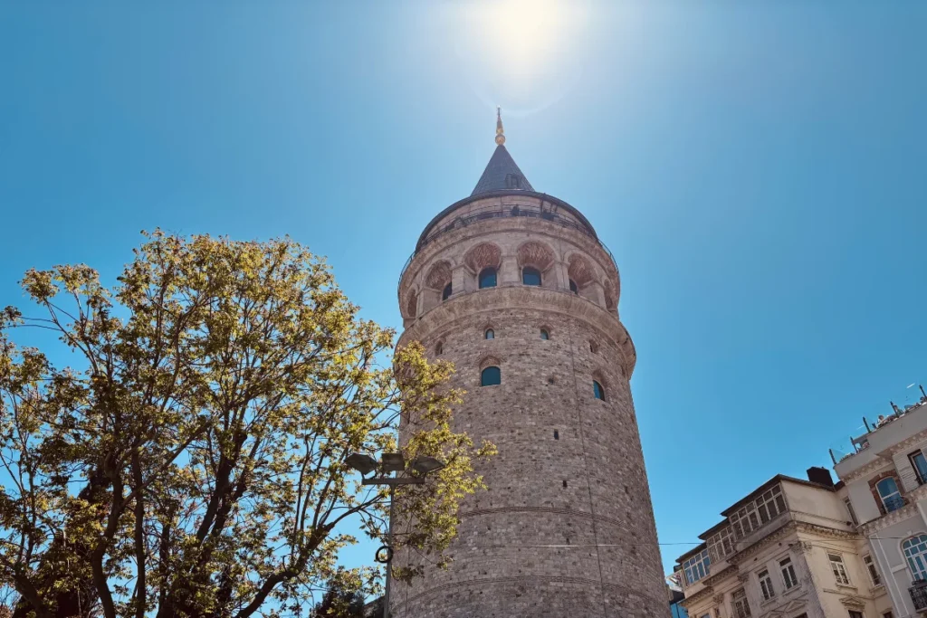 The historic stone Galata Tower rising above lush green trees in Istanbul under a bright blue sky.