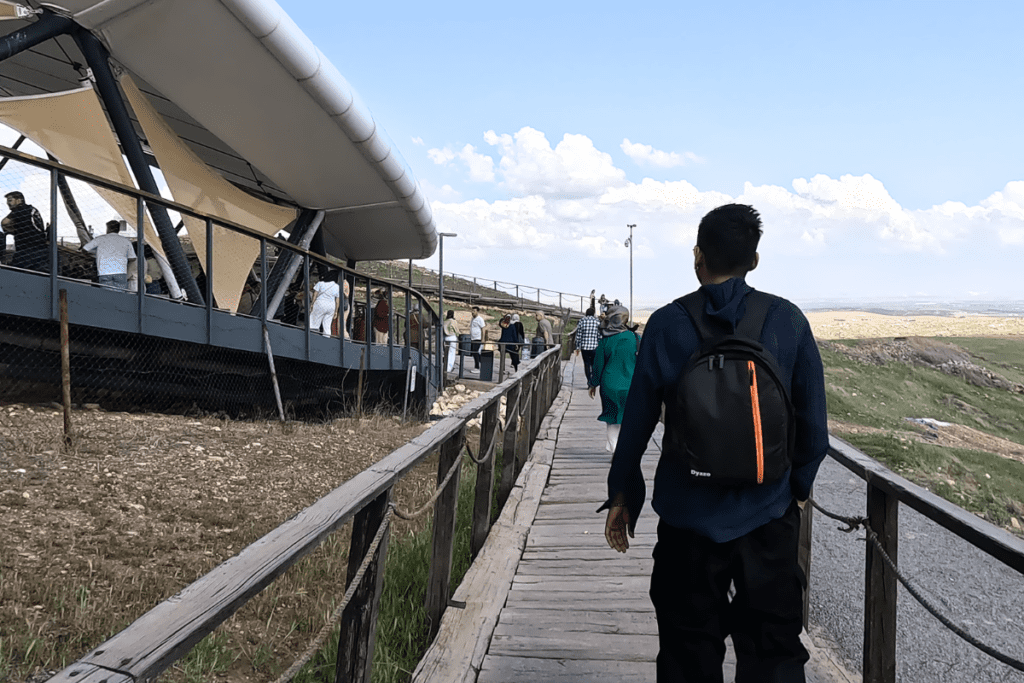 A rear view of a traveler walking along a modern wooden boardwalk leading toward the large canopy covering the Göbekli Tepe archaeological site.