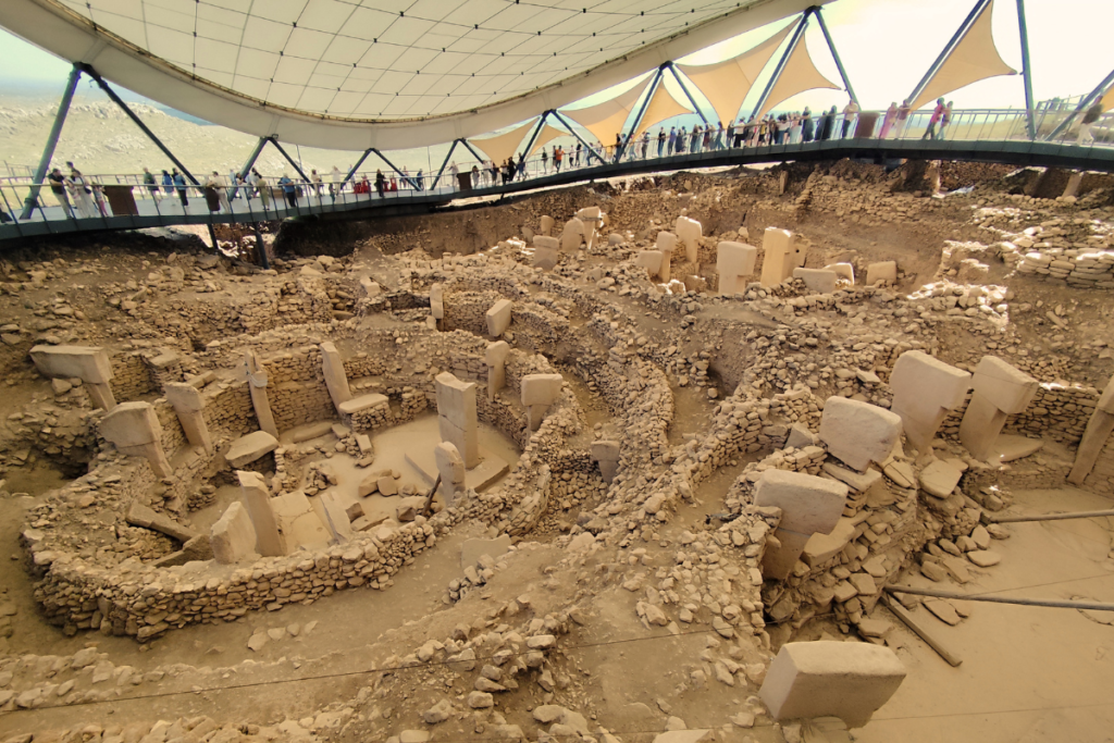 A wide panoramic shot of the archaeological complex at Göbekli Tepe. Multiple circular ancient stone ruins are visible inside a massive excavated valley, completely sheltered beneath a large, modern white protective roof structure with an elevated boardwalk for tourists.