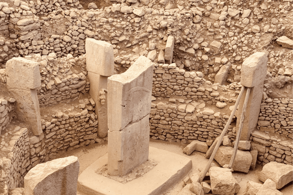 A high-angle, wide-set photograph looking down into a dry stone excavation pit. Several large, upright T-shaped limestone monoliths stand symmetrically within the curved, stacked stone retaining walls.