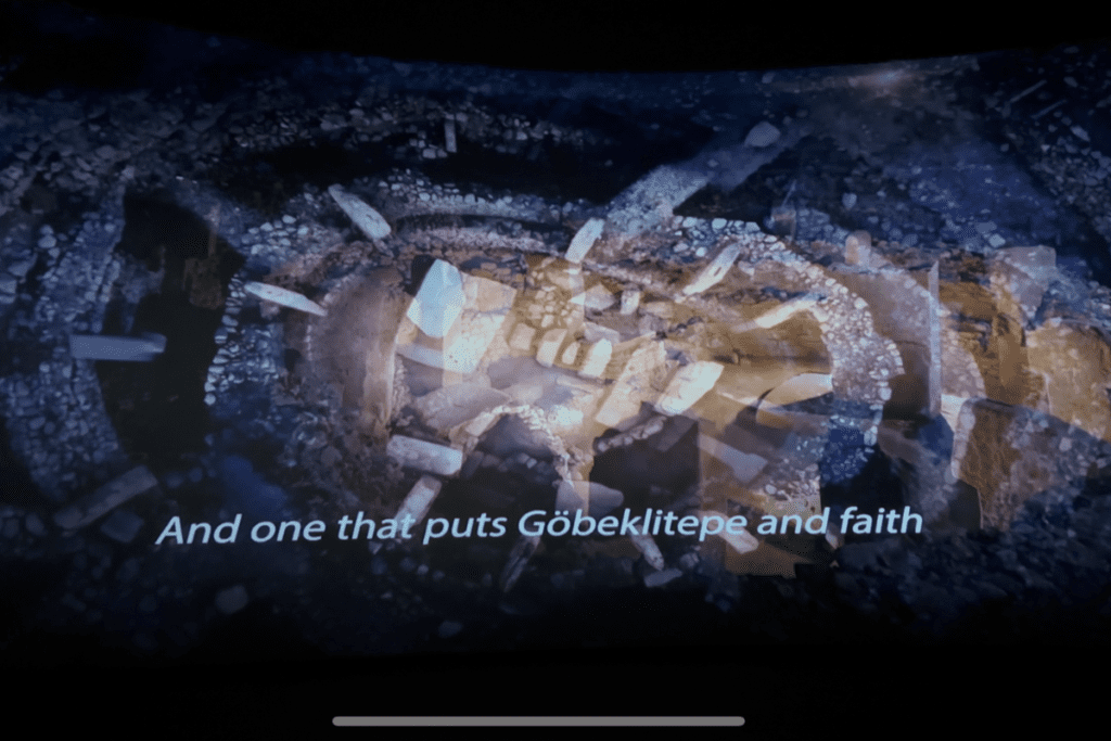 A dark museum exhibition screen displaying a glowing blue and white 3D digital projection of the circular stone enclosures, with the subtitle text "And one that puts Göbeklitepe and faith" overlaid at the bottom.