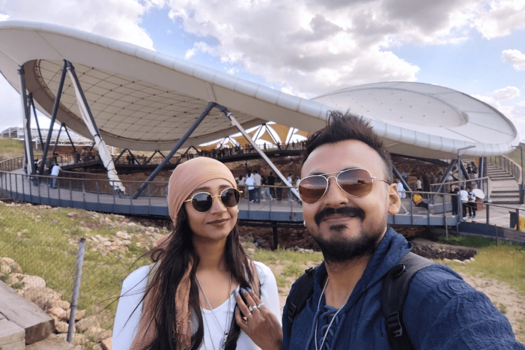 A happy, close-up outdoor photo of a married couple, Akash and Sumana, smiling together while wearing stylish sunglasses in front of Gobekli Tepe