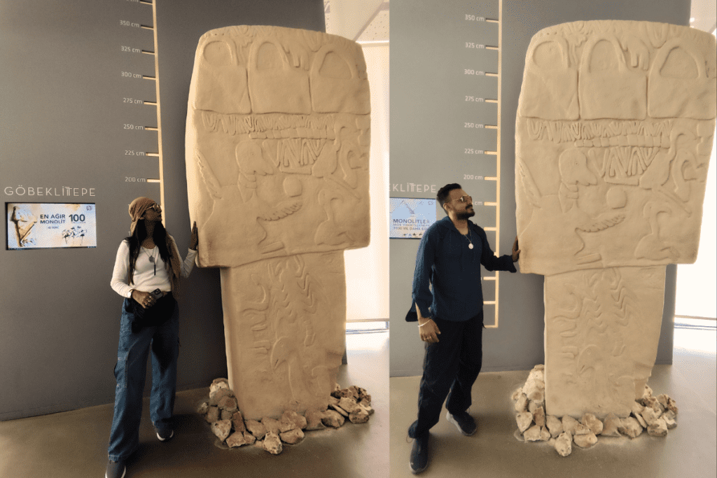 A side-by-side composite photo showing Sumana on the left and Akash on the right, each standing next to a massive, towering replica of an ancient T-shaped pillar inside a Gobekli Tepe museum gallery.